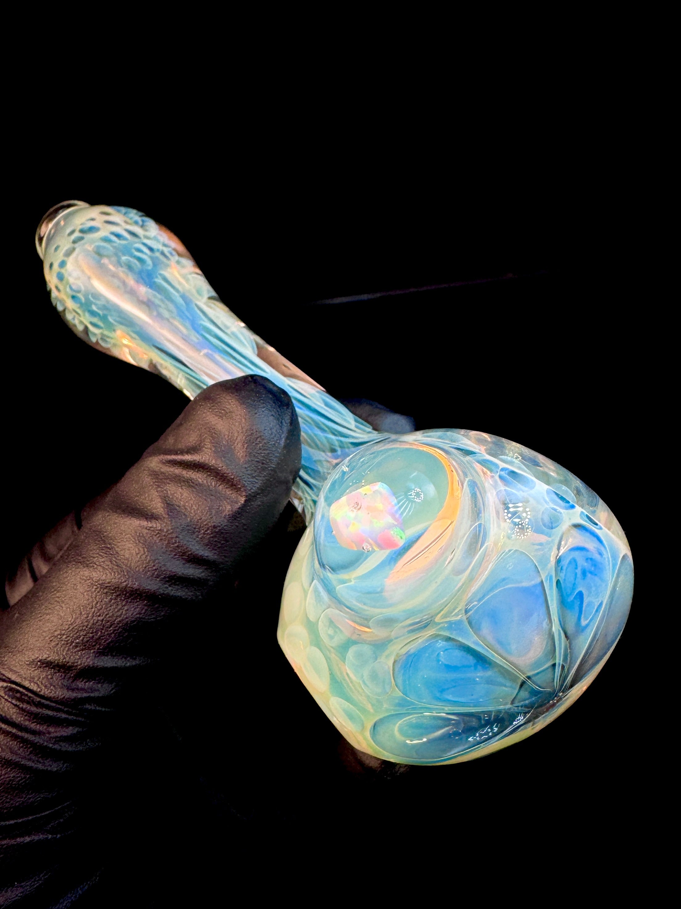 Glass pipe held by a gloved hand against a black background. The pipe has an opal and is fumed with silver.