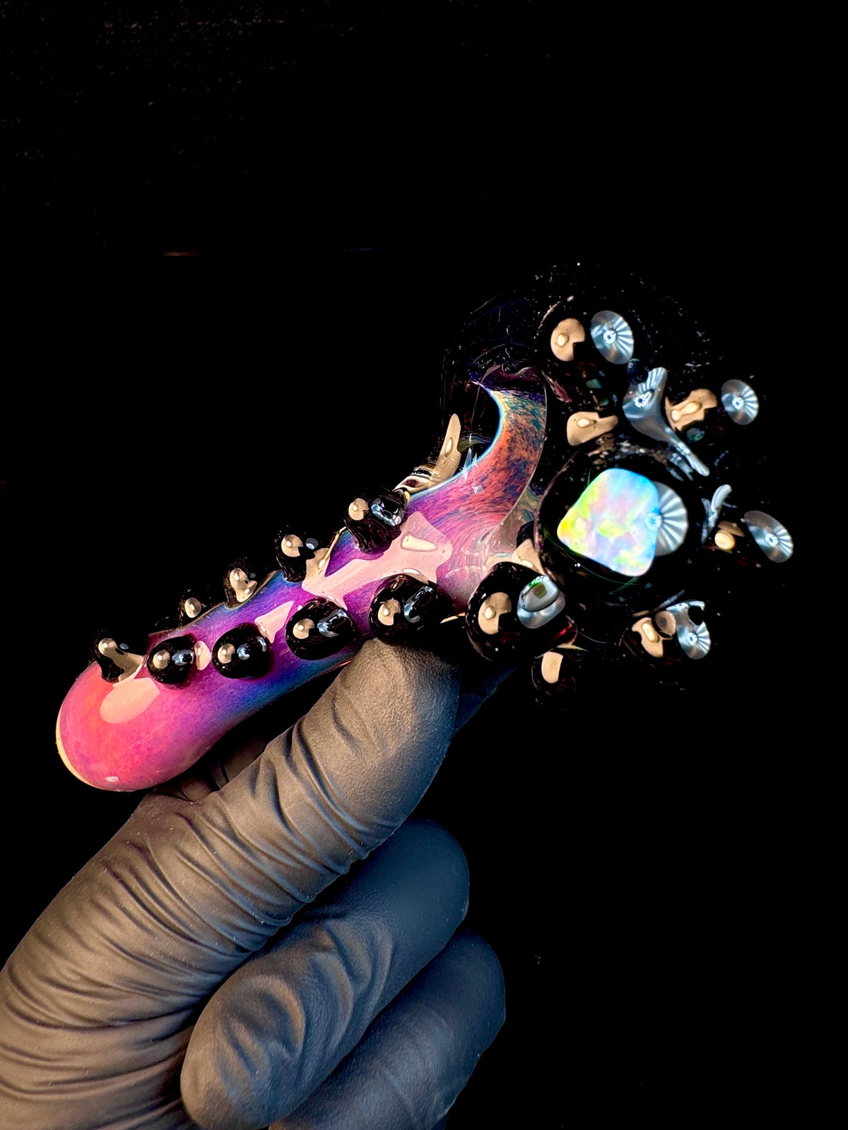 A hand holding a colorful glass pipe with black, pink, and purple hues, featuring a bulbous shape and small opal-like decorations.